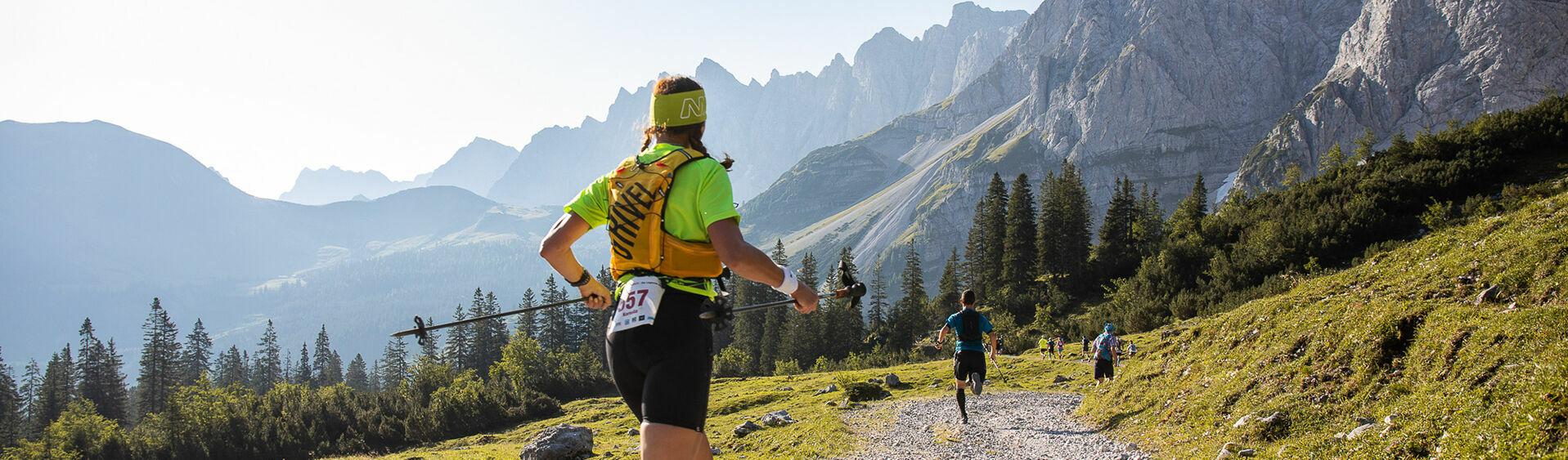 Ein paar Läufer des Karwendelmarschs laufen neben einer schönen Kulisse aus großen Bergen, grünen Wiesen und blauem Himmel.