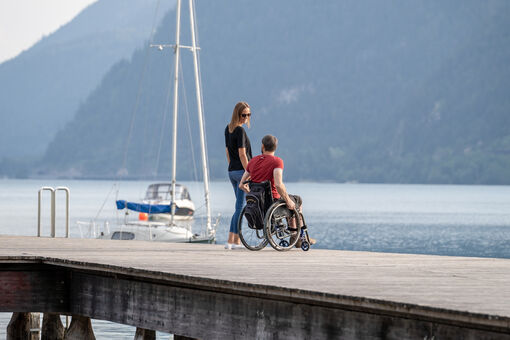 Ein Mann im Rollstuhl und eine Frau befinden sich auf dem Steg bei der Seeuferpromenade in Pertisau im Sommer. 