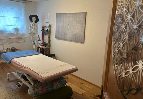 A bright therapy room features a massage table with a pink cover and a blue towel. A human skeleton model stands nearby, and various massage tools are displayed on a wall-mounted rack. Natural light filters in through a window with sheer curtains.