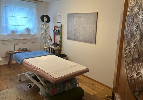 Das Bild zeigt einen Behandlungsraum mit einem Massagetisch, der mit einer blauen Decke bedeckt ist. An der Wand hängt ein großes Bild mit einem Rastermuster. Ein Skelettmodell steht neben dem Tisch, und es gibt einige therapeutische Utensilien in der Nähe.