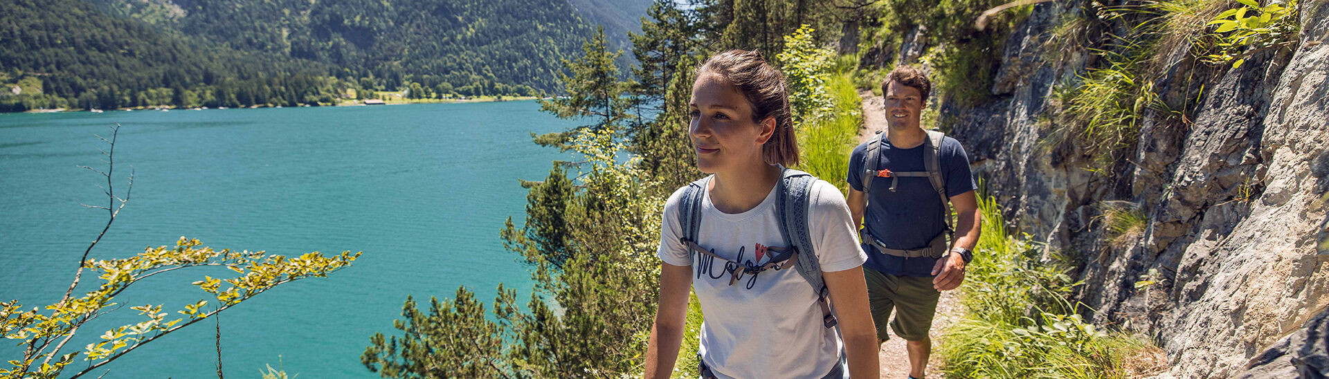 Ein Pärchen spaziert auf einem schmalen steinernen Weg neben dem türkisblauem Achensee Richtung Gaisalm.