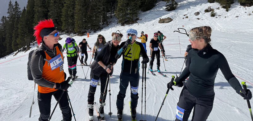 A group of skiers is gathered on a snowy mountain slope, preparing for a race. Some participants wear playful costumes, including a person with a bright red mohawk and another with antler-like accessories. The atmosphere is lively and festive, showcasing a spirit of fun and competition.