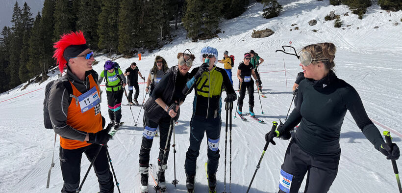 Eine Gruppe von Skitourengeher steht auf schneebedecktem Gelände, bereit für einen Wettkampf. Einige Teilnehmer tragen auffällige Kostüme, wie eine Frau mit Brille. Die Stimmung ist fröhlich und unbeschwert.