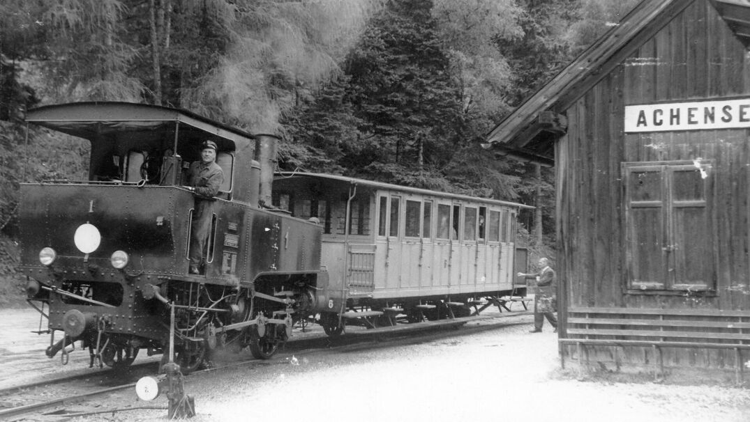 Historische Aufnahme der Achenseebahn mit Dampflokomotive Historische Schwarz-Weiß-Aufnahme der Achenseebahn mit Dampflokomotive und Personenwagen an einer Haltestelle. Ein Lokführer steht auf der Lokomotive, daneben befindet sich ein hölzernes Bahnhofsgebäude mit der Aufschrift "Achensee".