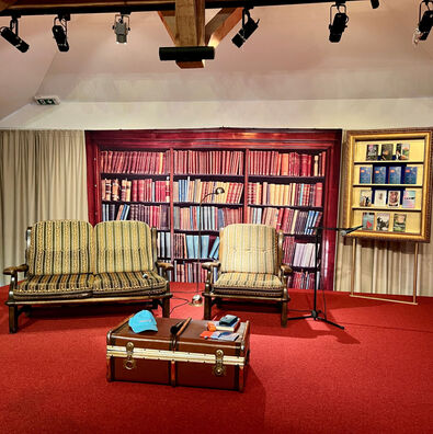 Altes Widum in Achenkirch Der Raum zeigt zwei gestreifte Sessel vor einer Bücherwand mit vielen bunten Buchrücken. Neben den Sesseln steht ein Koffer und ein Mikrofon. Im Hintergrund hängen Bilderrahmen mit Buchcovern. Der Boden ist rot, und es gibt eine Leuchter mit Kerzen.