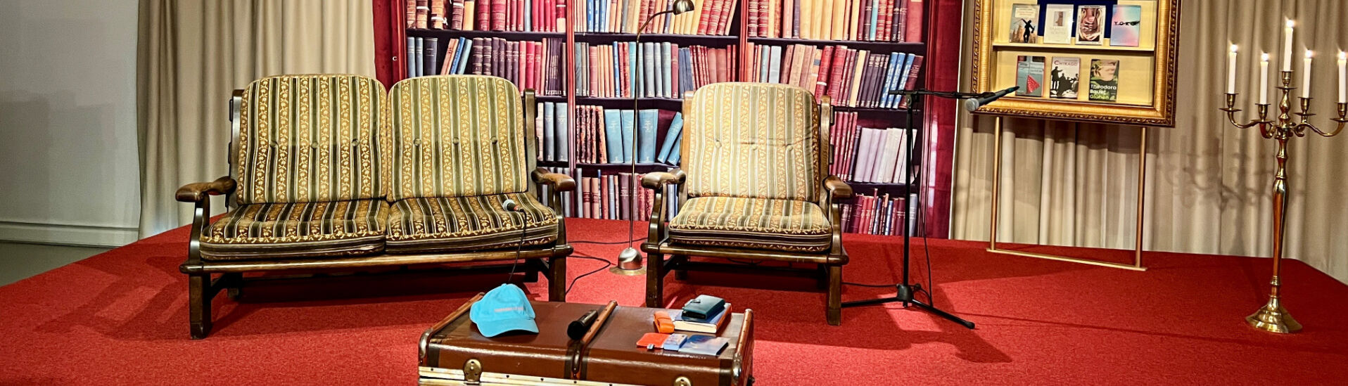 Altes Widum in Achenkirch Der Raum zeigt zwei gestreifte Sessel vor einer Bücherwand mit vielen bunten Buchrücken. Neben den Sesseln steht ein Koffer und ein Mikrofon. Im Hintergrund hängen Bilderrahmen mit Buchcovern. Der Boden ist rot, und es gibt eine Leuchter mit Kerzen.