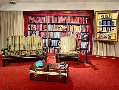 Der Raum zeigt zwei gestreifte Sessel vor einer Bücherwand mit vielen bunten Buchrücken. Neben den Sesseln steht ein Koffer und ein Mikrofon. Im Hintergrund hängen Bilderrahmen mit Buchcovern. Der Boden ist rot, und es gibt eine Leuchter mit Kerzen.