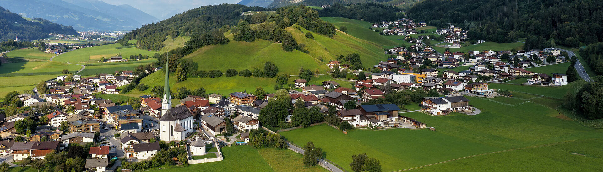 Blick auf Wiesing am Fuße des Rofangebirges. Der Ort ist von saftig grünen Wiesen und sanften Hügeln umgeben, während sich im Hintergrund die Gipfel des Rofan erheben.