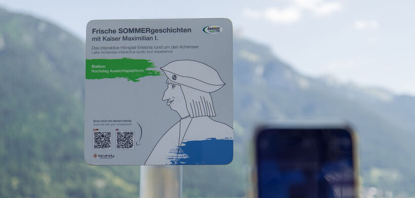 A sign promoting an interactive audio experience titled "Fresh Summer Stories with Emperor Maximilian I." It indicates the location as "Hochsteg Viewing Platform" and includes instructions to scan a QR code with a smartphone for more information. The background features mountains and a lake.