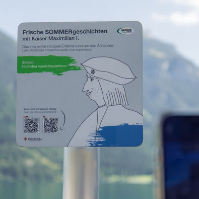 Hörspiel-Erlebnis am Achensee mit Kaiser Maximilian I. Ein Informationsschild am Aussichtspunkt Hochsteg am Achensee. Es bewirbt eine interaktive Hörspiel-Tour mit Kaiser Maximilian I. und enthält Anweisungen zum Scannen eines QR-Codes, um mehr über die Geschichte und die Umgebung zu erfahren.