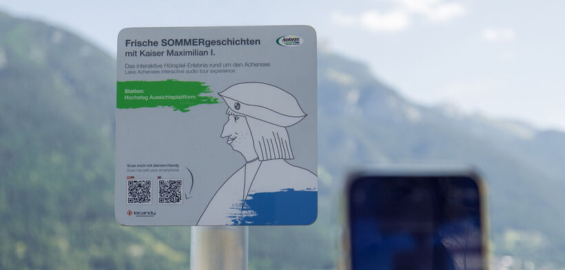 Ein Informationsschild am Aussichtspunkt Hochsteg am Achensee. Es bewirbt eine interaktive Hörspiel-Tour mit Kaiser Maximilian I. und enthält Anweisungen zum Scannen eines QR-Codes, um mehr über die Geschichte und die Umgebung zu erfahren.