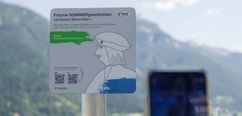 Ein Informationsschild am Aussichtspunkt Hochsteg am Achensee. Es bewirbt eine interaktive Hörspiel-Tour mit Kaiser Maximilian I. und enthält Anweisungen zum Scannen eines QR-Codes, um mehr über die Geschichte und die Umgebung zu erfahren.