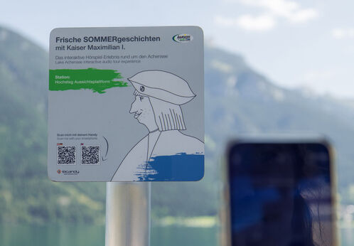 Digitales Hörspiel-Erlebnis mit Kaiser Max Ein Informationsschild am Aussichtspunkt Hochsteg am Achensee. Es bewirbt eine interaktive Hörspiel-Tour mit Kaiser Maximilian I. und enthält Anweisungen zum Scannen eines QR-Codes, um mehr über die Geschichte und die Umgebung zu erfahren.