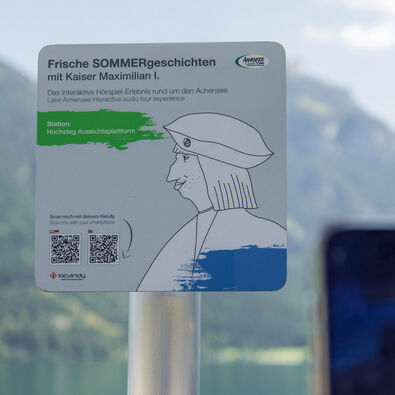 A sign promoting an interactive audio experience titled "Fresh Summer Stories with Emperor Maximilian I." It indicates the location as "Hochsteg Viewing Platform" and includes instructions to scan a QR code with a smartphone for more information. The background features mountains and a lake.