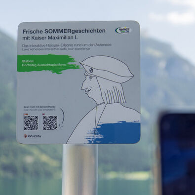Ein Informationsschild am Aussichtspunkt Hochsteg am Achensee. Es bewirbt eine interaktive Hörspiel-Tour mit Kaiser Maximilian I. und enthält Anweisungen zum Scannen eines QR-Codes, um mehr über die Geschichte und die Umgebung zu erfahren.