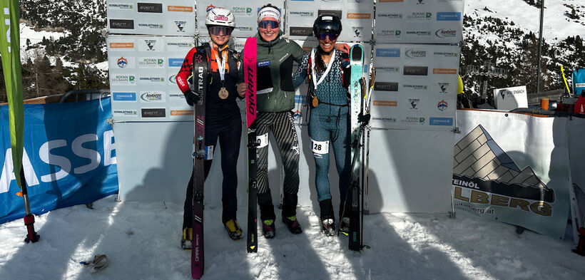 Three female skiers stand together on snow, each holding skis and wearing medals around their necks. They are smiling, celebrating their achievements in a skiing competition.