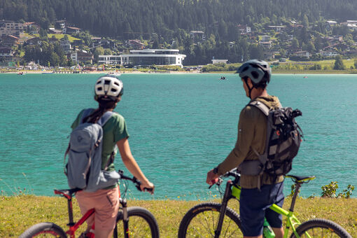 A couple explores the lakeshore in Maurach am Achensee by bike. In the background of this photo is the Atoll Achensee.