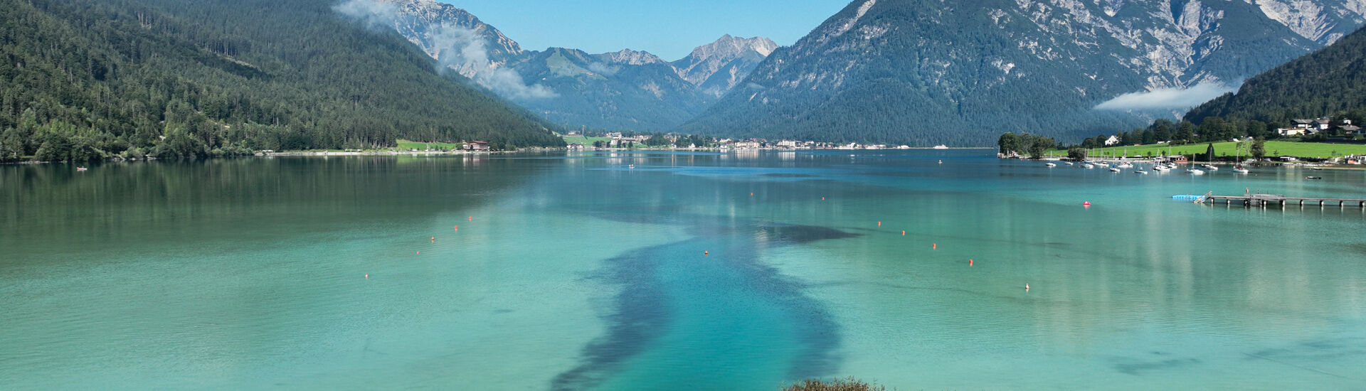 Wankratzbach am Südufer des Achensees Eine ruhige Landschaft mit dem glitzernden, türkisfarbenen Achensee, umgeben von hohen Bergen und Wäldern. Im Vordergrund wächst das Schilf.