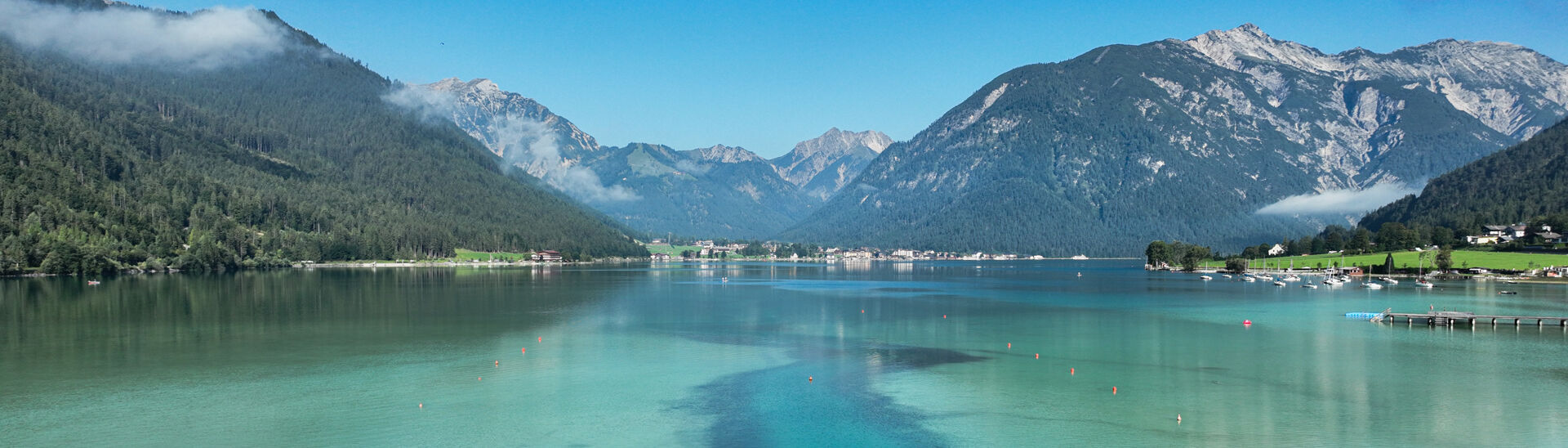 The Lake Achensee is bordered by lush green mountains under a clear blue sky. The water displays shades of turquoise and hints of reflection from the surrounding landscape. A narrow channel leads through patches of reeds, suggesting a peaceful and natural setting.