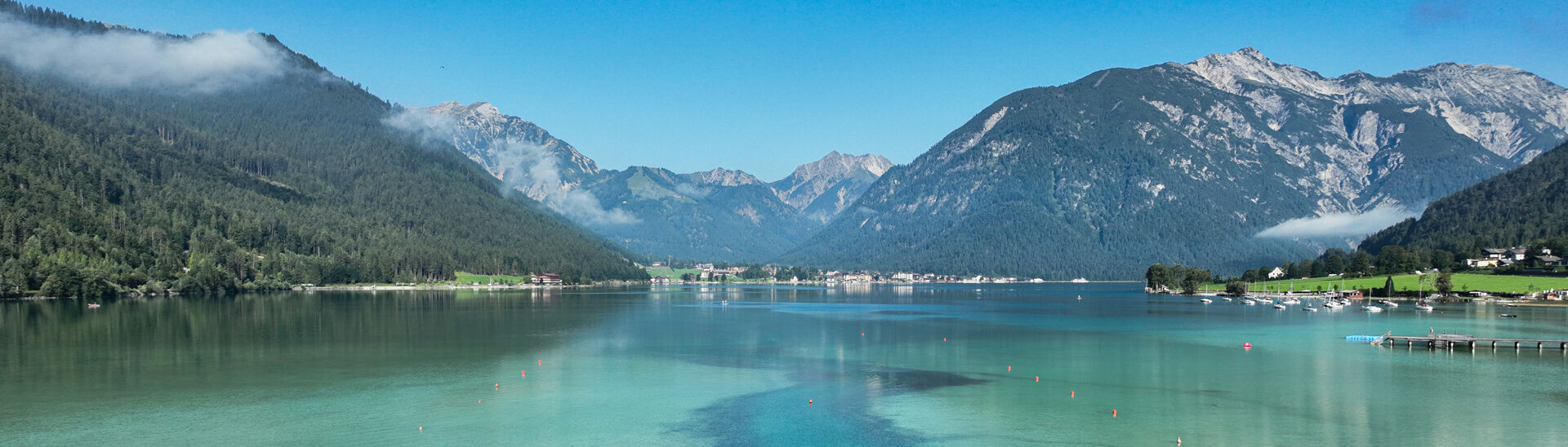 Eine ruhige Landschaft mit dem glitzernden, türkisfarbenen Achensee, umgeben von hohen Bergen und Wäldern. Im Vordergrund wächst das Schilf.