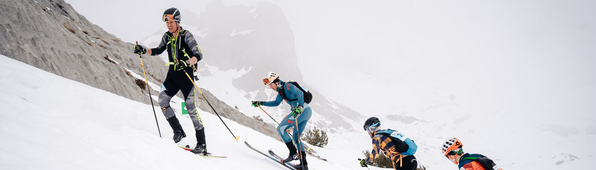 Vier Skifahrer erklimmen einen schneebedeckten Hang im Rofangebirge. Sie tragen technische Ski-Ausrüstung und Skischuhe. Die Umgebung ist gebirgig und in Wolken gehüllt, was die Sicht einschränkt. Alle Sportler sind konzentriert und in Bewegung, während sie den anspruchsvollen Aufstieg bewältigen.