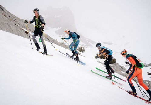 Four skiers ascend a snowy slope, each using ski poles for support. They wear various brightly colored ski gear as they navigate the steep terrain. The environment is mountainous and overcast, creating a wintry scene.