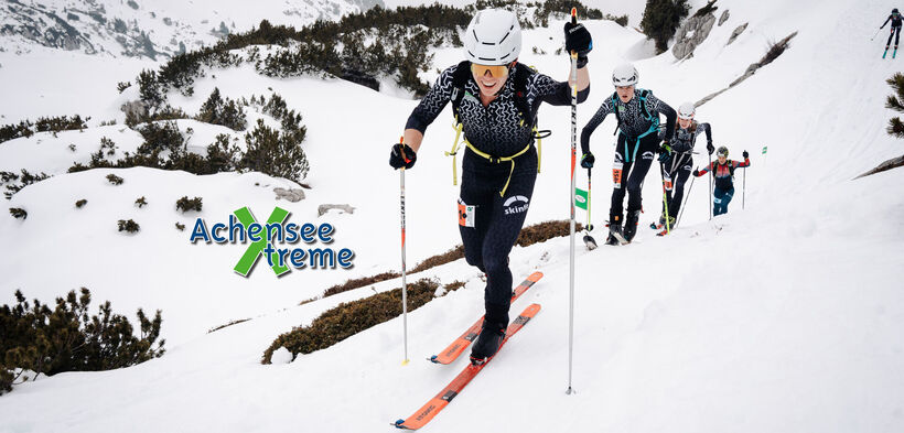 A group of skiers ascends a snowy mountain, navigating through a winter landscape with patches of vegetation. The lead skier demonstrates determination while using ski poles, and the background shows additional skiers making their way up the slope in a cold, cloudy setting.