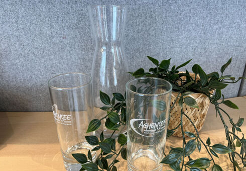 Your Achensee welcome gift The image features two glass drinking tumblers and a tall glass carafe. In the background, there is a woven basket containing decorative greenery. The items are arranged on a wooden surface against a gray backdrop.
