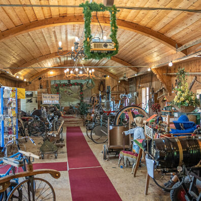 Das Bild zeigt das Innere der Achenseer Museums- und Erlebniswelt mit Holzdecke. Es sind verschiedene alte Maschinen und Werkzeuge ausgestellt, darunter Fahrräder und landwirtschaftliche Geräte. Ein roter Teppich verläuft durch den Raum, der mit Pflanzen und dekorativen Elementen geschmückt ist.