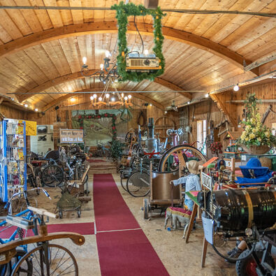 The image depicts a wide interior of a rustic museum filled with vintage machinery and artifacts. A red carpet runs down the center, flanked by various historical items, including bicycles and engines, under a wooden ceiling with warm lighting and decorations, giving a nostalgic atmosphere.