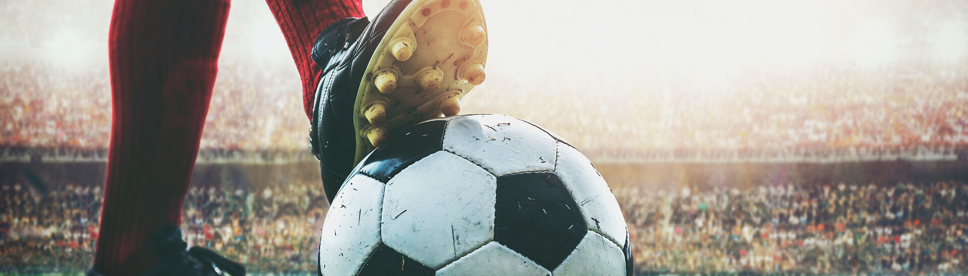 Nahaufnahme eines Fußballs auf einem grünen Spielfeld. Ein Spieler mit roten Stutzen und schwarzen Fußballschuhen hat den Fuß auf den Ball gestellt. Im Hintergrund ist ein voll besetztes Stadion mit unscharfen Zuschauerrängen zu sehen.