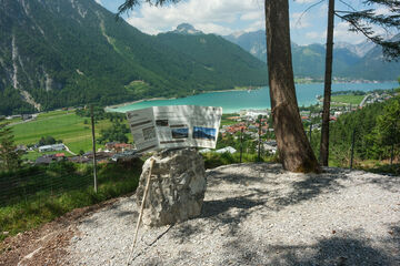 Aussichtspunkt oberhalb des Achensees mit einer Informationstafel auf einem Steinblock. Von der Plattform aus fällt der Blick auf den türkisblauen See, umliegende Ortschaften, grüne Wiesen und bewaldete Berghänge.
