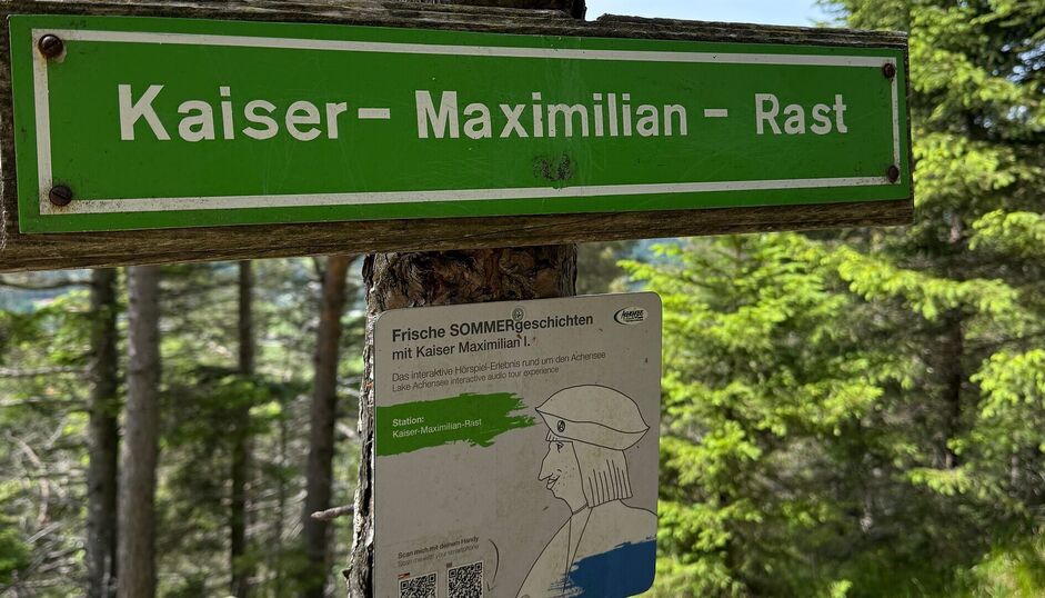 Nahaufnahme der Beschilderung an der Kaiser-Maximilian-Rast im Wald über dem Achensee. Unter dem grünen Wegschild befindet sich eine Infotafel zu den „Frischen Sommergeschichten“ mit Kaiser Maximilian.