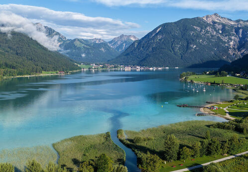 Drohnenaufnahme über den türkisblauen Achensee im Sommer. Das glitzernde Wasser und der sattgrüne Schilfgürtel prägen das Landschaftsbild. /// A drone shot of turquoise-blue Lake Achensee in summer, where glistening waters and a lush green belt of reeds characterize the landscape.