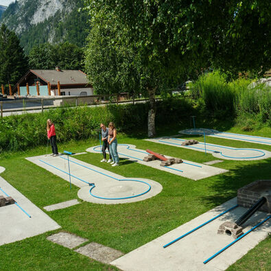 Auf dem Bild ist ein Minigolfplatz in Maurach in einem Garten zu sehen. Drei Personen stehen an verschiedenen Bahnen. Die Bahnen sind in einem grünen Bereich angeordnet, umgeben von Bäumen und Gras. Im Hintergrund sind einige Gebäude sichtbar, die eine alpine Umgebung schaffen.