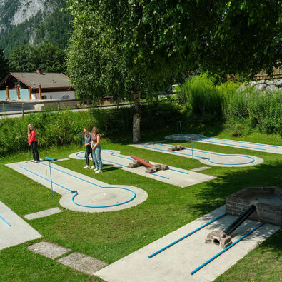A mini-golf course set in a green outdoor area in Maurach features multiple putt paths designed with blue outlines. Three individuals are engaged in the game, with one about to take a shot while the others observe. Trees and a building are visible in the background.