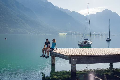 A couple exploring the lakeside promenade and the wooden pier in Pertisau am Achensee, backdropped by the Ebner Joch and several sailboats.