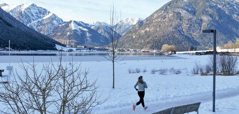 Eine Trailrunnerin läuft im Winter am Achensee entlang und im Hintergrund sieht man das schneebedeckte Karwendel und die umliegende Berglandschaft. Neben ihr liegt der Achensee mit ruhiger Wasseroberfläche.