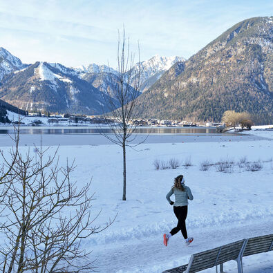 Eine Trailrunnerin läuft im Winter am Achensee entlang und im Hintergrund sieht man das schneebedeckte Karwendel und die umliegende Berglandschaft. Neben ihr liegt der Achensee mit ruhiger Wasseroberfläche.