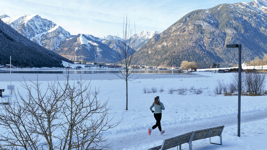 Eine Trailrunnerin läuft im Winter am Achensee entlang und im Hintergrund sieht man das schneebedeckte Karwendel und die umliegende Berglandschaft. Neben ihr liegt der Achensee mit ruhiger Wasseroberfläche.