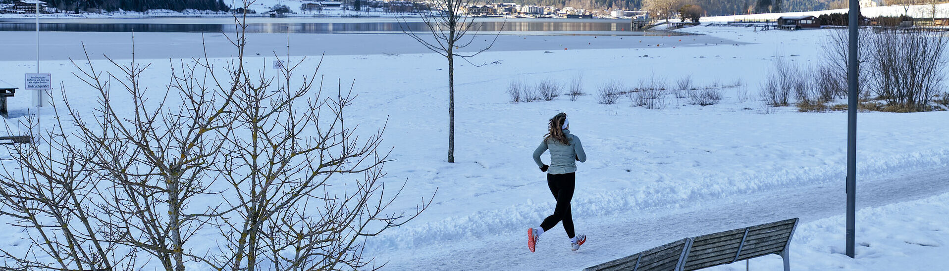 Eine Trailrunnerin läuft im Winter am Achensee entlang und im Hintergrund sieht man das schneebedeckte Karwendel und die umliegende Berglandschaft. Neben ihr liegt der Achensee mit ruhiger Wasseroberfläche.