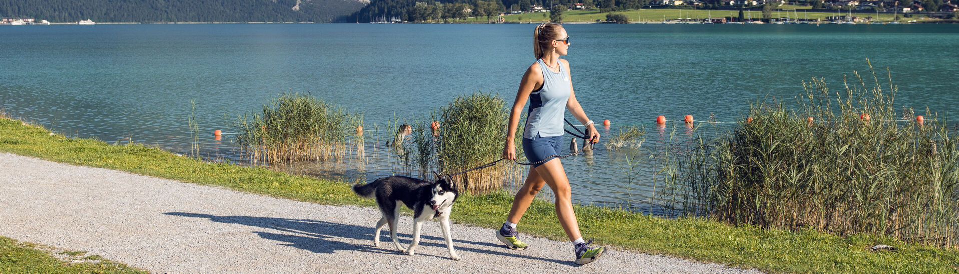 A woman walks alongside lake Achensee on a sunny day, accompanied by a black and white dog. The surrounding landscape features mountains and greenery, creating a peaceful outdoor scene. The path is gravel, bordered by grassy areas and gentle water.