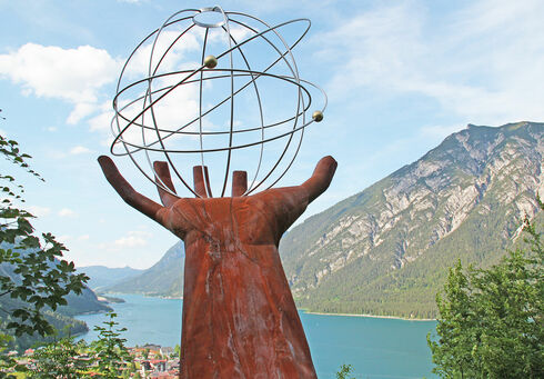 Link öffnen Eine Skulptur einer Hand die einen eisernen durchsichtigen Planeten auf dem Besinnungsweg am in Pertisau hält.