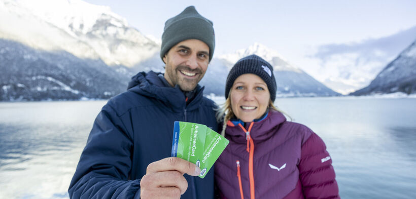Ein Paar hält die AchenseeCard in die Kamera. Im Hintergrund die herrliche Winterlandschaft am Achensee.