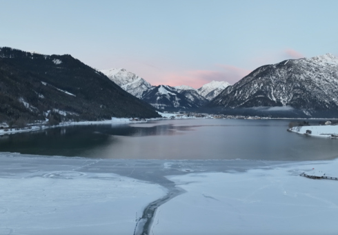 Der Blick zeigt die ruhige Winterlandschaft am Südufer des Achensees mit einem teilweise zugefrorenen See, umgeben von schneebedeckten Bergen. Der Himmel ist blau mit zarten rosa Wolken.