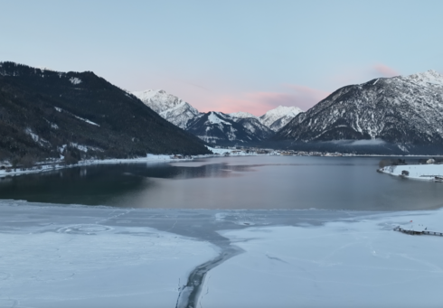 The view shows the tranquil winter landscape on the southern shore of Lake Achensee with a partially frozen lake surrounded by snow-covered mountains. The sky is blue with delicate pink clouds.