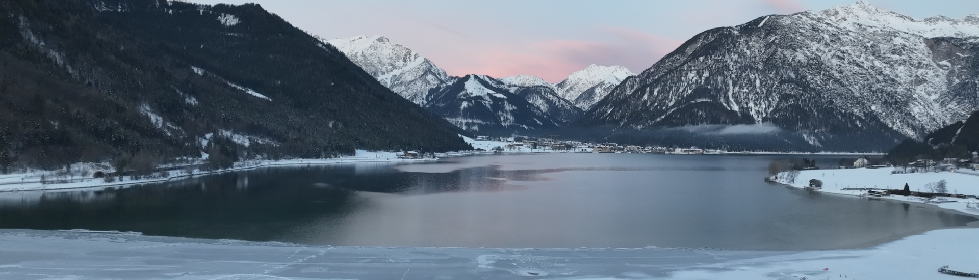 Der Blick zeigt die ruhige Winterlandschaft am Südufer des Achensees mit einem teilweise zugefrorenen See, umgeben von schneebedeckten Bergen. Der Himmel ist blau mit zarten rosa Wolken.