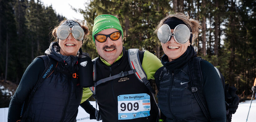 Three cheerful people stand on the event route of the Achensee Ski Touring Fun and smile into the camera.