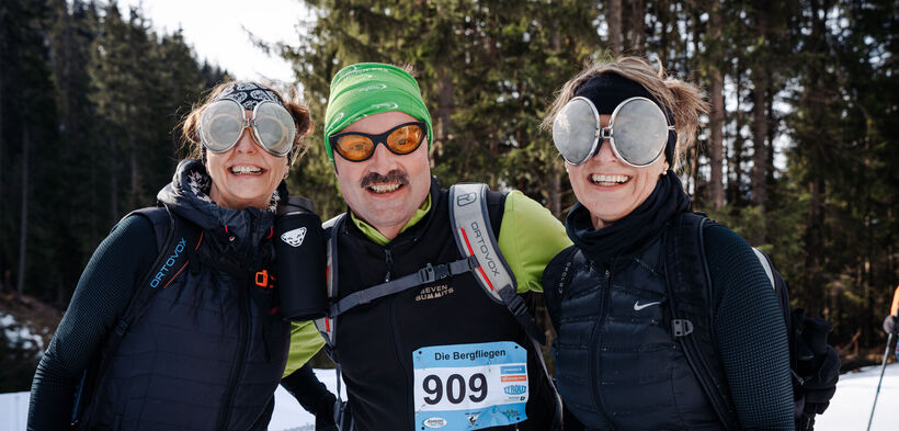 Drei fröhliche Personen stehen auf der Eventstrecke des Achensee Skitourenspaß und lächeln in die Kamera. Sie tragen auffällige Sonnenbrillen und Sportbekleidung. 