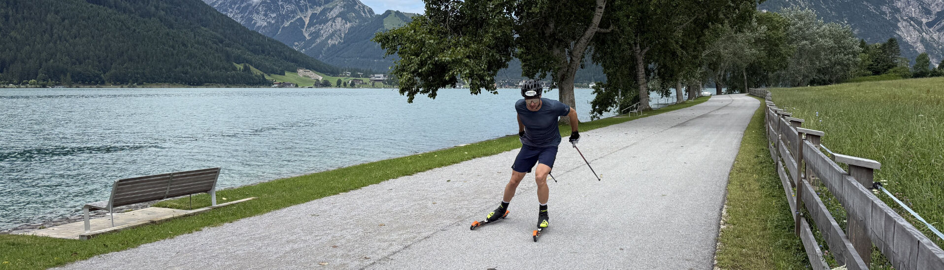 Ein Mann fährt mit Rollski auf einem asphaltierten Weg entlang eines Achensees. Er trägt einen Helm, Handschuhe und Sportbekleidung und nutzt Stöcke zum Antrieb.